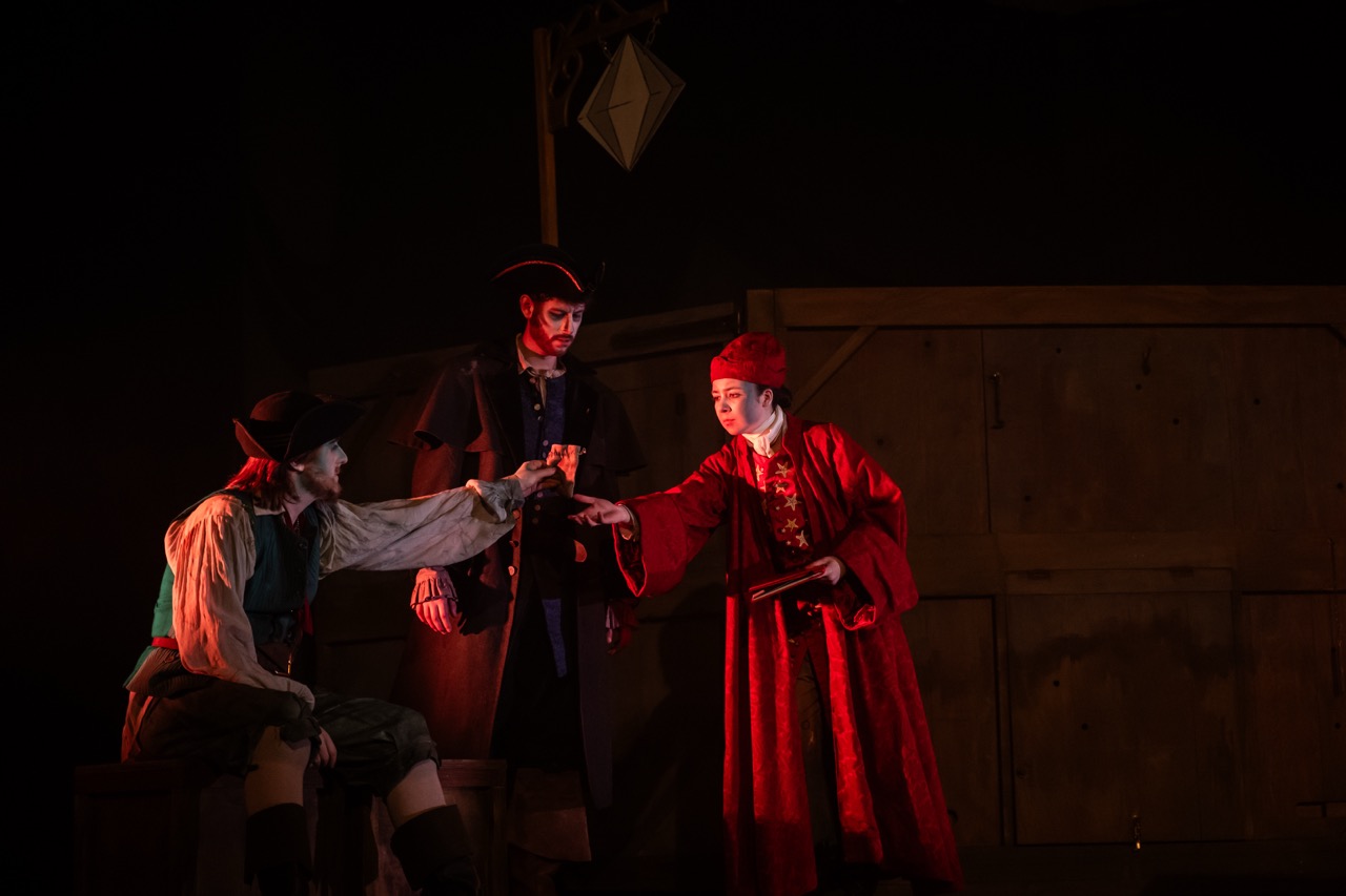 Three actors perform on stage in a theatrical setting. One actor dressed in a red robe holds something out towards another actor in a green and white outfit, while the third actor in a dark coat observes. The background features wooden panels.