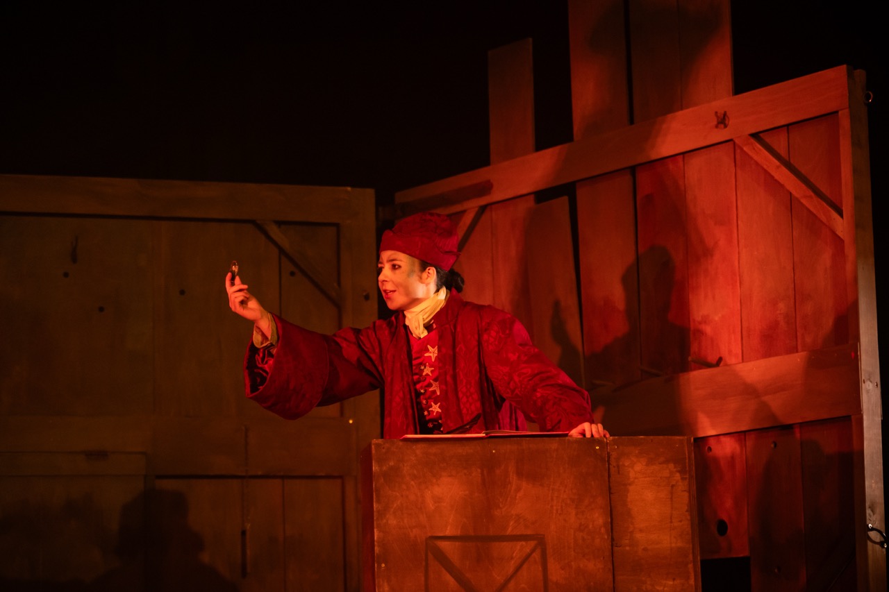 A performer wearing a red costume and hat gestures while standing behind a wooden stage set. The background features wooden panels with warm lighting casting shadows.
