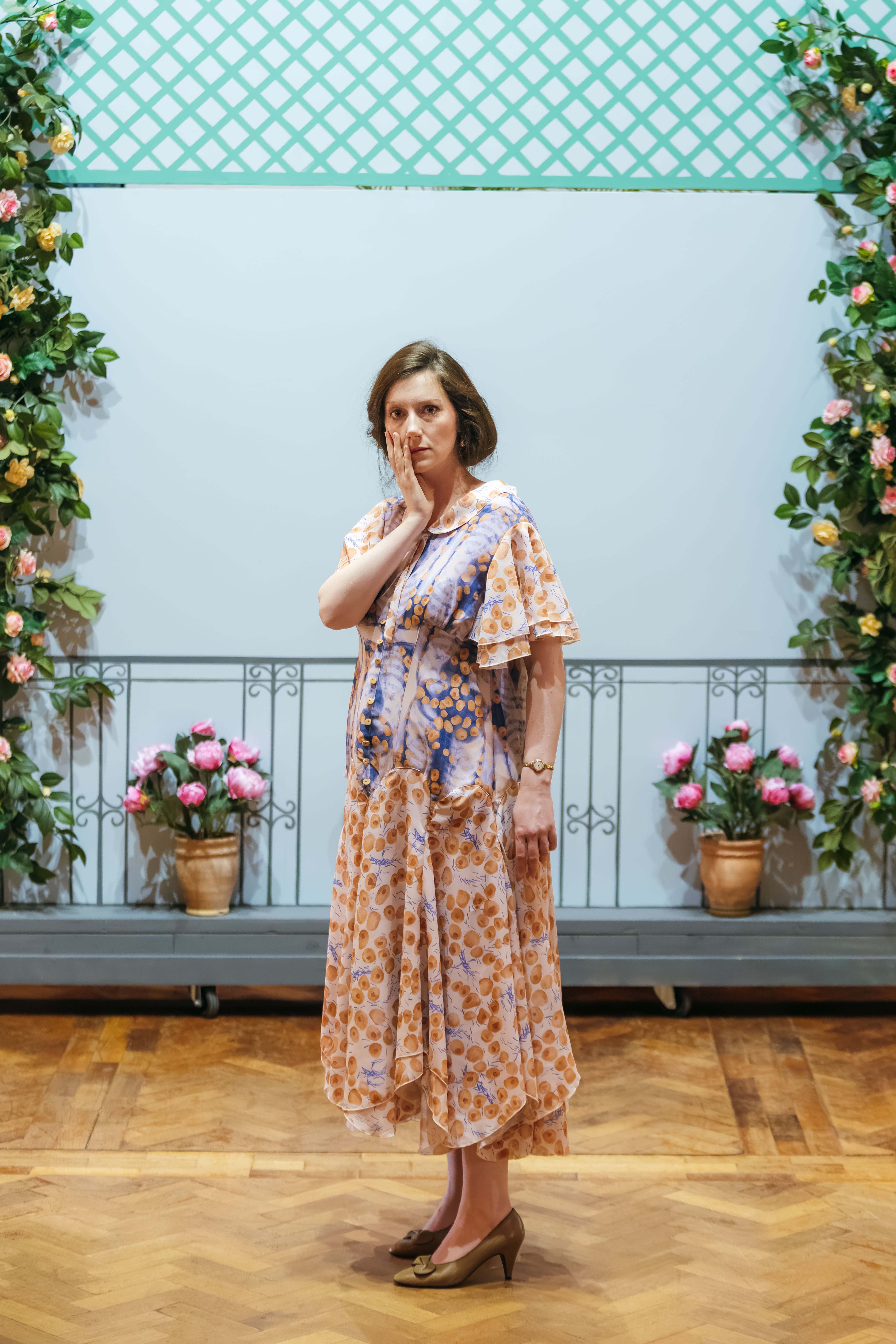 A woman wearing a floral patterned dress stands with her hand near her face in a posed manner. The background features green lattice and pink flowers arranged in pots, with a wooden floor beneath her.