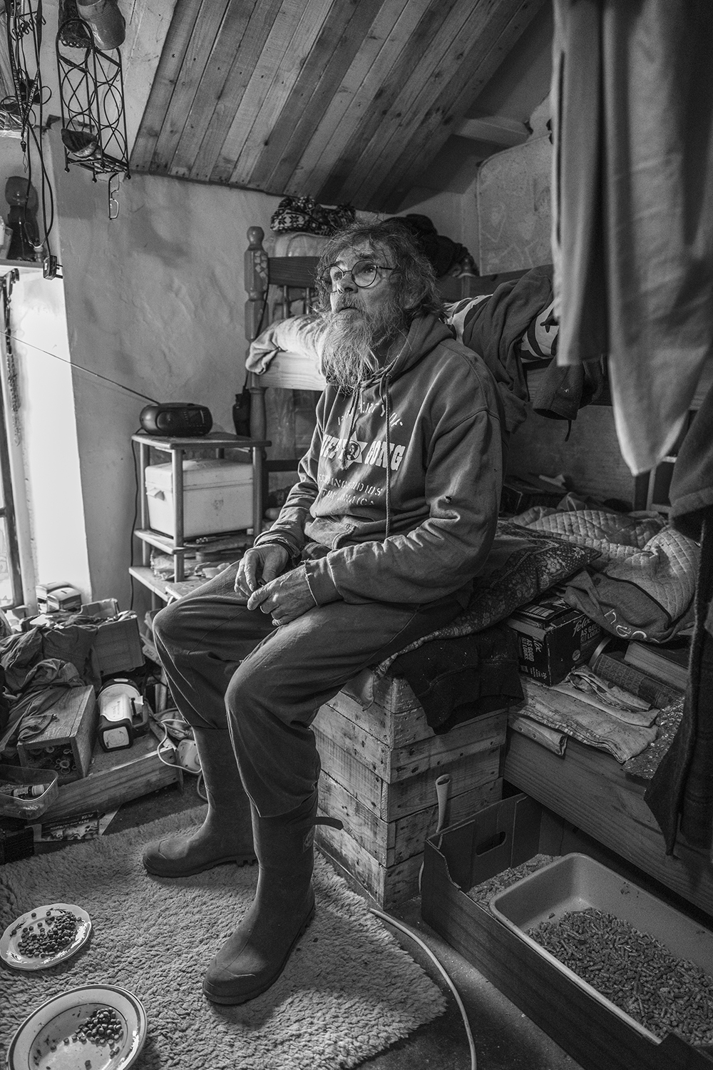 Elderly bearded man with glasses sits on wooden crate in cluttered shed interior. Black and white image with exposed ceiling beams.