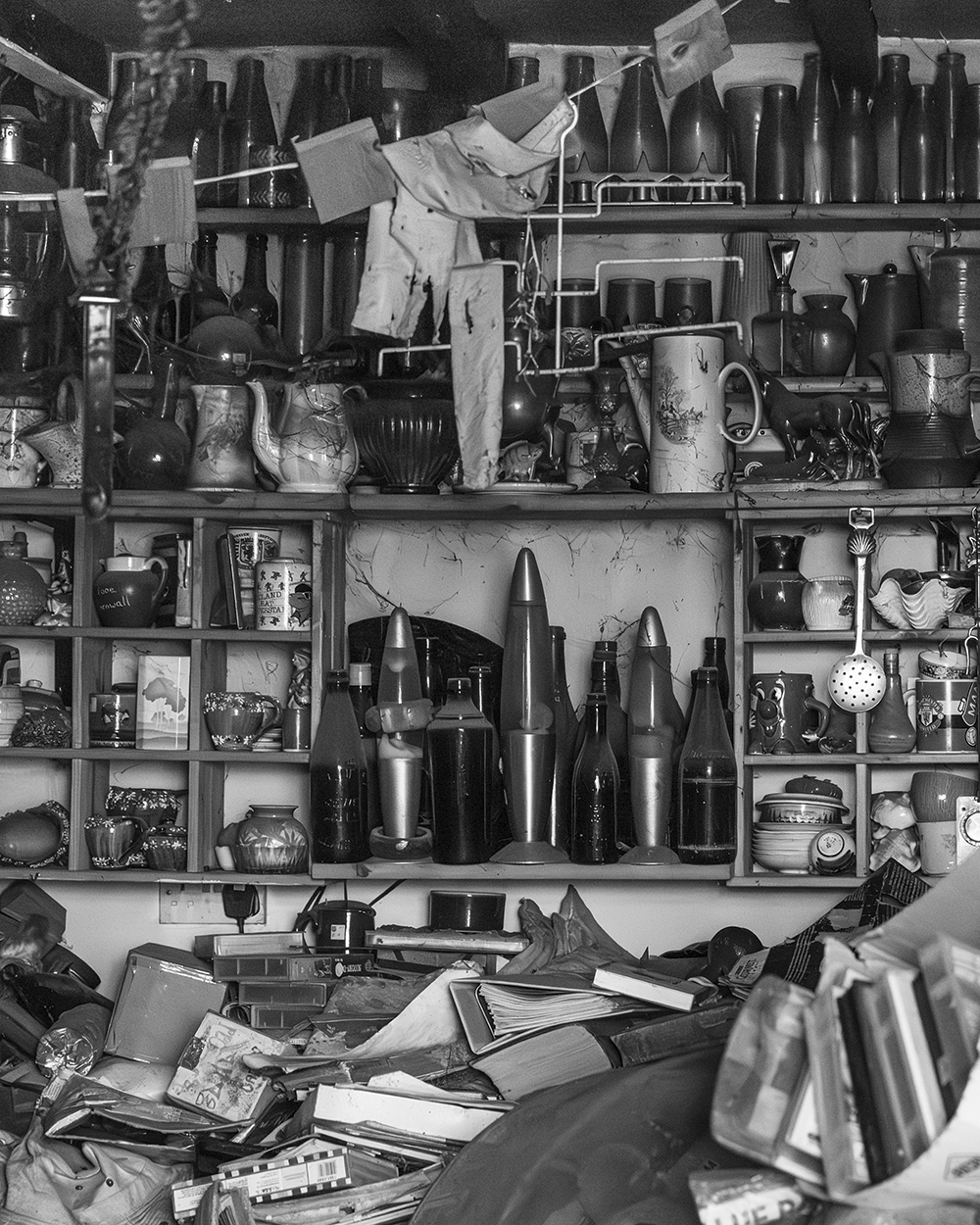 Black and white image showing cluttered shelves filled with bottles, jars, containers and various objects with papers scattered below.
