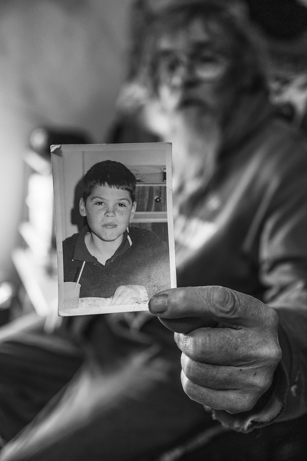 Black and white image of hands holding vintage photograph of young boy; blurred figure in background.