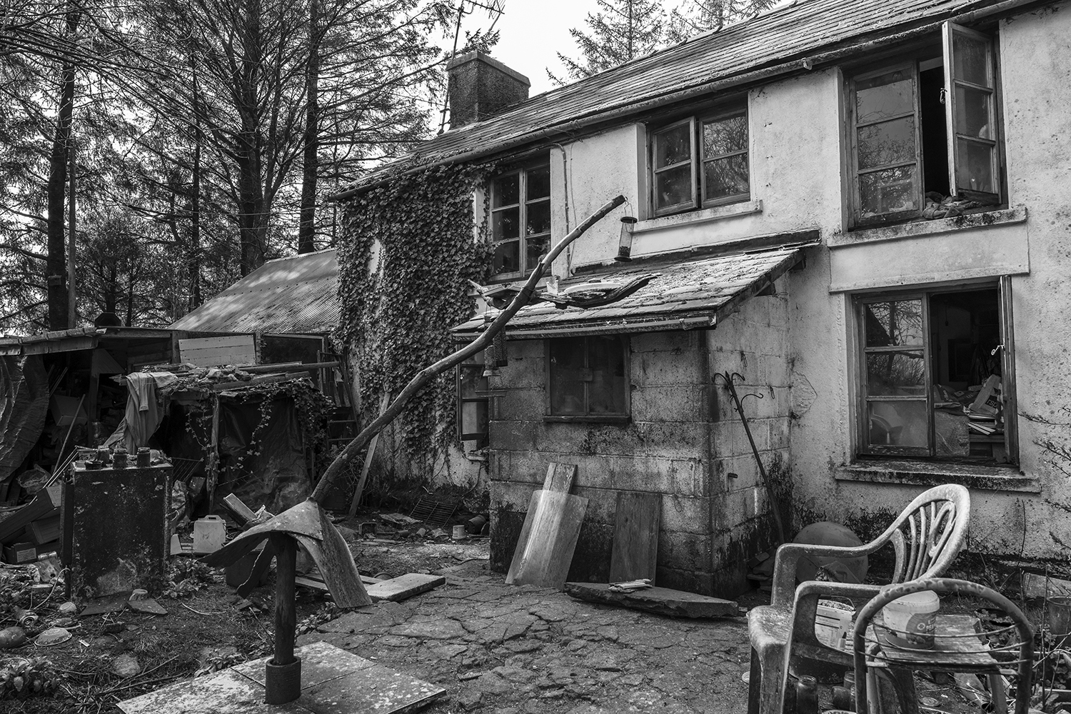 Derelict two-storey house with broken windows, external stairs, scattered debris and plastic chair in foreground. Black and white image.