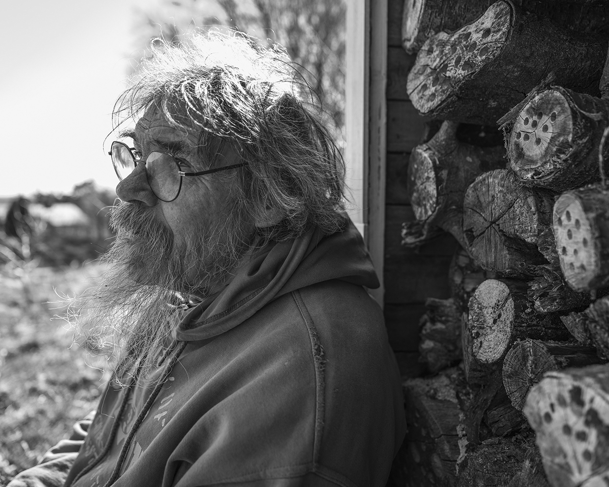 Elderly bearded man with glasses and hooded jacket in profile, beside stacked logs. Black and white photography.