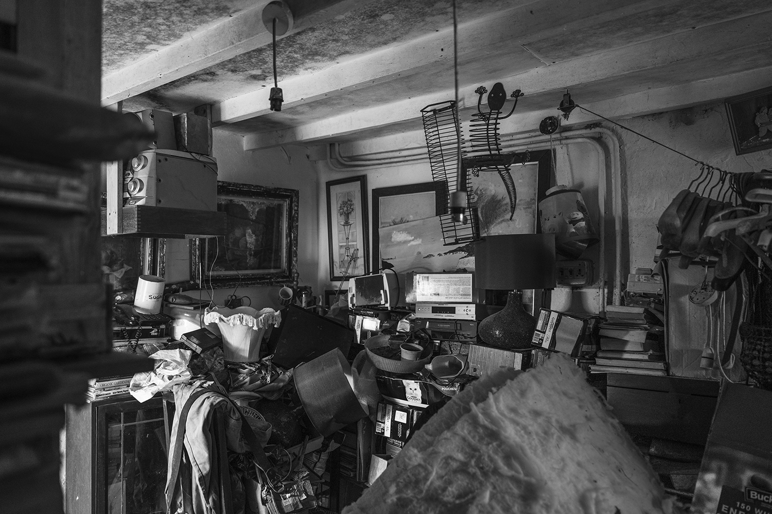 Black and white image of cluttered interior room with exposed ceiling beams, various equipment, books, and furniture scattered throughout.