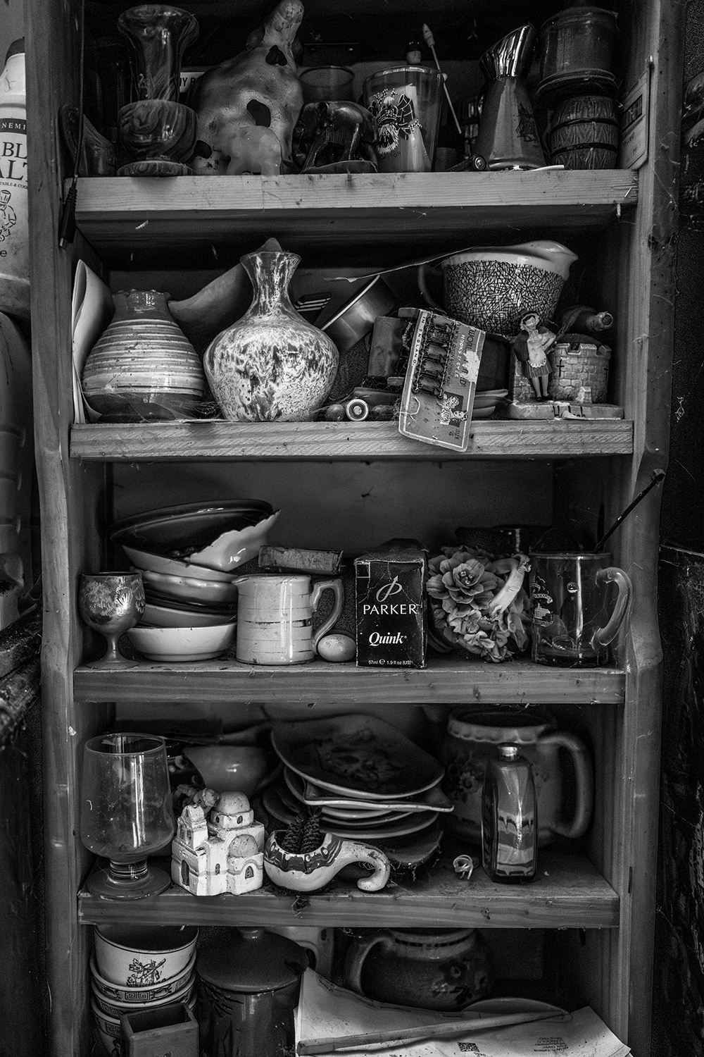 Black and white image of wooden shelves densely packed with ceramic vessels, pottery, bowls, jugs, and miscellaneous objects.
