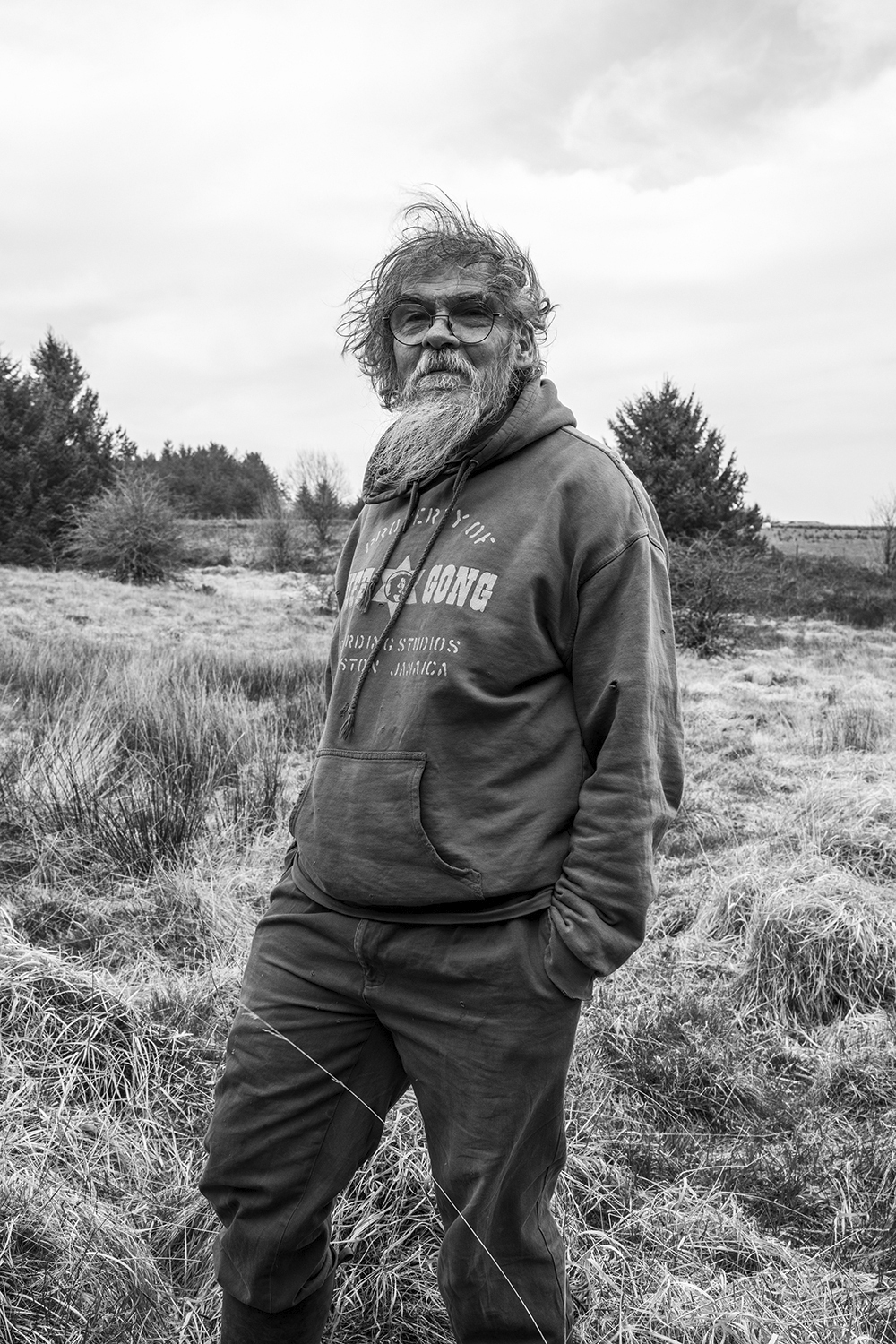 Elderly man with long grey hair and beard wearing hoodie stands in grassy field with trees and cloudy sky behind him.