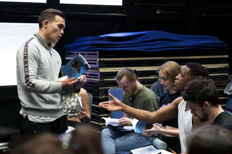 BA (Hons) Acting students rehearsing; four people are shown sitting, another person is stood up in front of them holding a book.