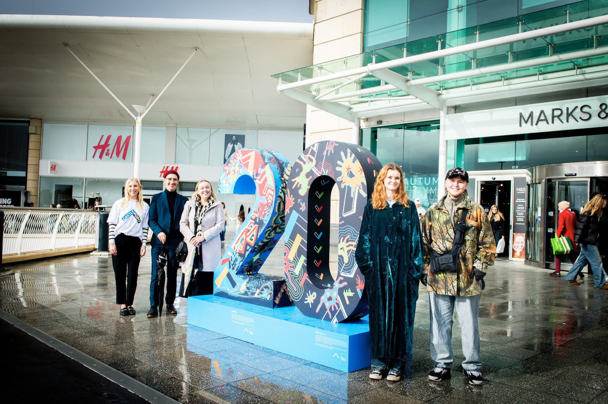 A large 3D number 20 standing on a pedestal outside Castlepoint Shopping Centre. AUB staff stand to the left, two AUB BA (Hons) Illustration students stand to the right, and pose for a photo.