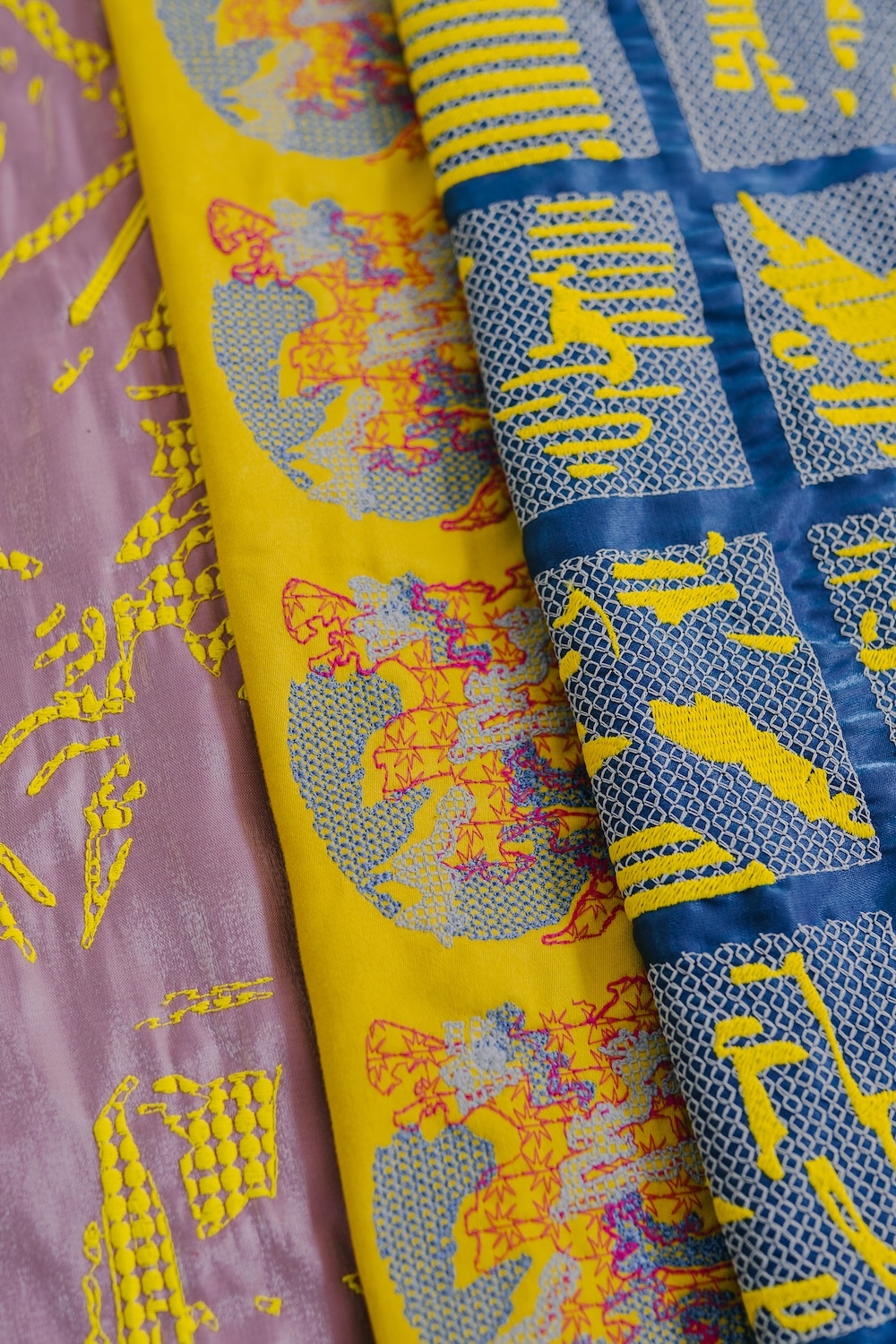 Colourful fabric scarves with yellow, blue and pink patterns featuring geometric shapes, dots and abstract designs in layered arrangement.