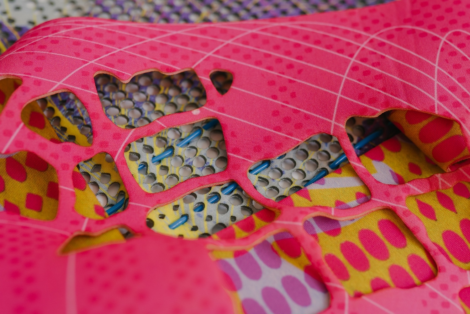 Pink mesh fabric with grid pattern overlaying multicoloured printed material featuring rows of circular dots in blue, yellow, and grey.