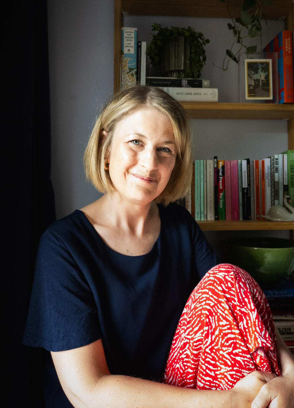 Woman with blonde bob haircut in navy top and red patterned trousers, smiling whilst seated beside wooden bookshelves filled with books.