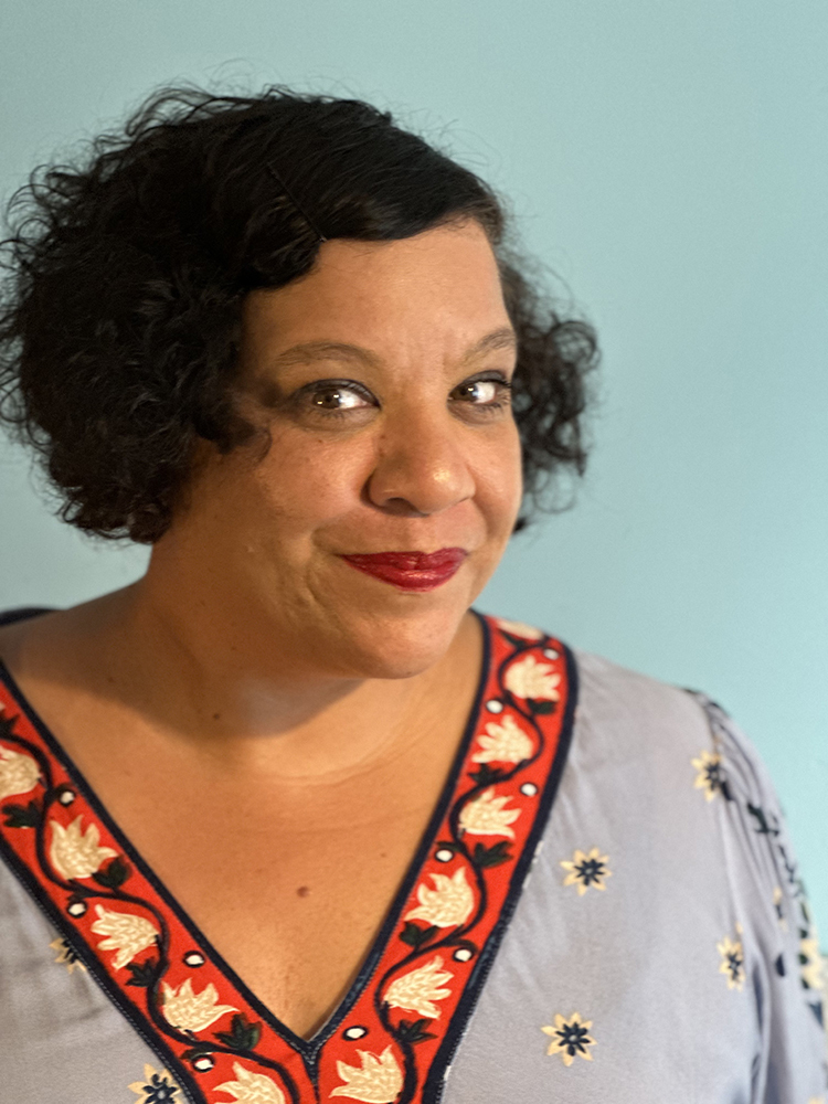 Woman with short curly dark hair wearing red lipstick and white top with red floral embroidered neckline against light blue background