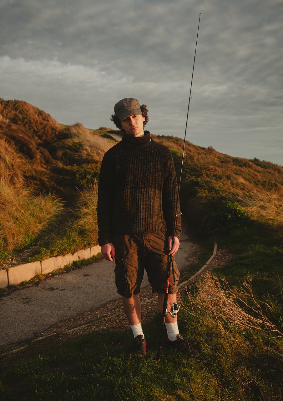 A young man stands on a path beside grassy hills, holding a fishing rod. He wears a dark knitted jumper, shorts, white socks, and black shoes, with a grey cap on his head. The sky is cloudy and the lighting is warm.