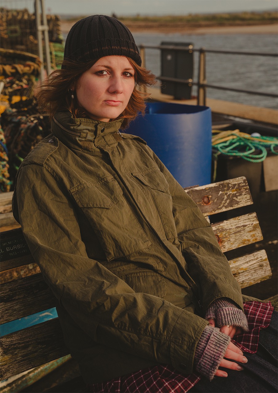 A woman with shoulder-length brown hair sits on a wooden bench wearing an olive green jacket and a black knitted hat. Behind her is a blue container and some coils of rope, with a body of water visible in the background.