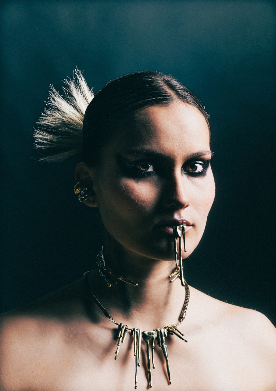A close-up portrait of a woman with sleek hair styled upwards, wearing a bold necklace and earrings. Metallic elements are integrated into her lower lip, creating a striking visual effect against a dark background.