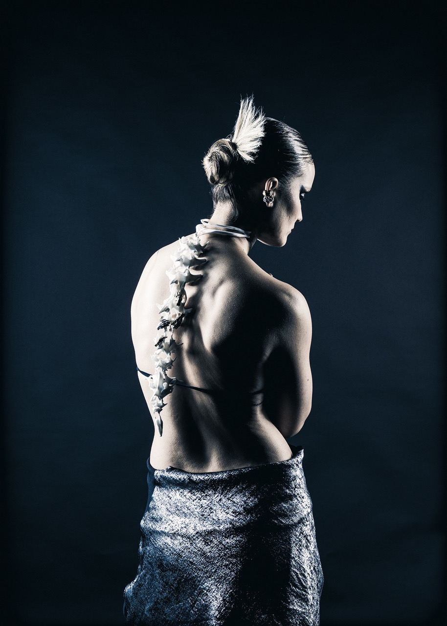 A woman stands with her back to the viewer, showcasing a decorative arrangement resembling a spine along her back. She has her hair tied up and is draped in a textured fabric that covers the lower part of her body, against a dark background.