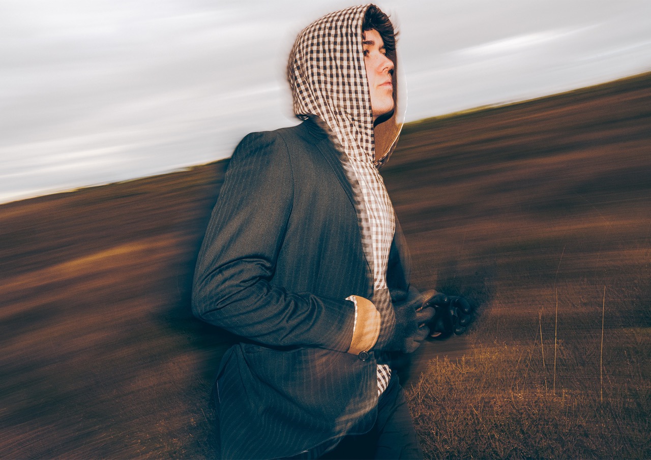 A young man wearing a striped suit jacket and a checkered hooded top walks against a blurred background of a grassy field and cloudy sky. The motion blur suggests speed or movement.