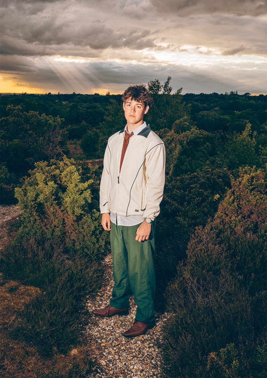 A young man stands amidst shrubbery, wearing a light grey jacket over a brown sweater and green trousers. He looks directly at the camera, and the background features a dramatic sky with beams of light breaking through the clouds.