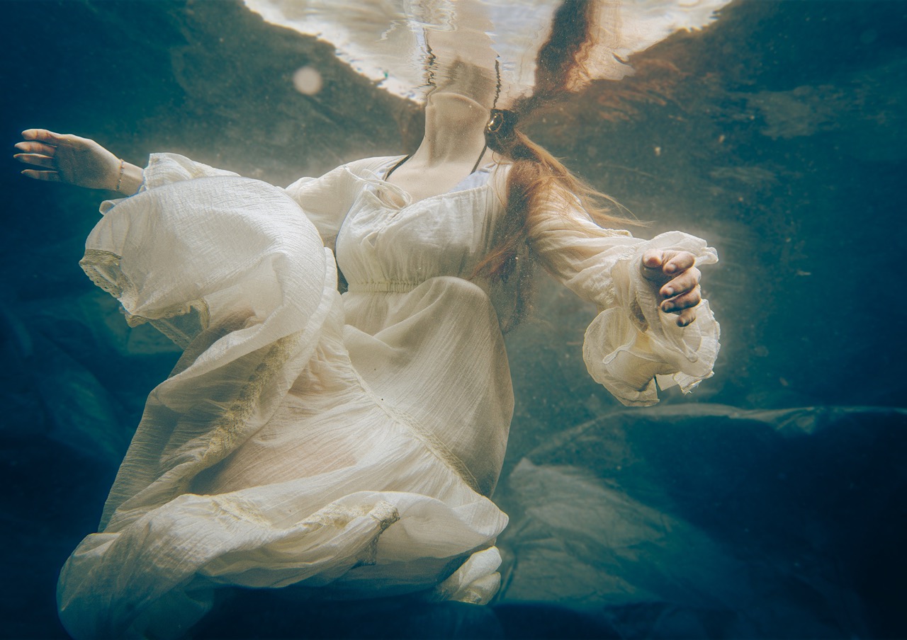 A figure in a flowing white dress is submerged in water, appearing to float gracefully. The image shows the dress billowing around the figure and captures the calmness of the underwater environment.
