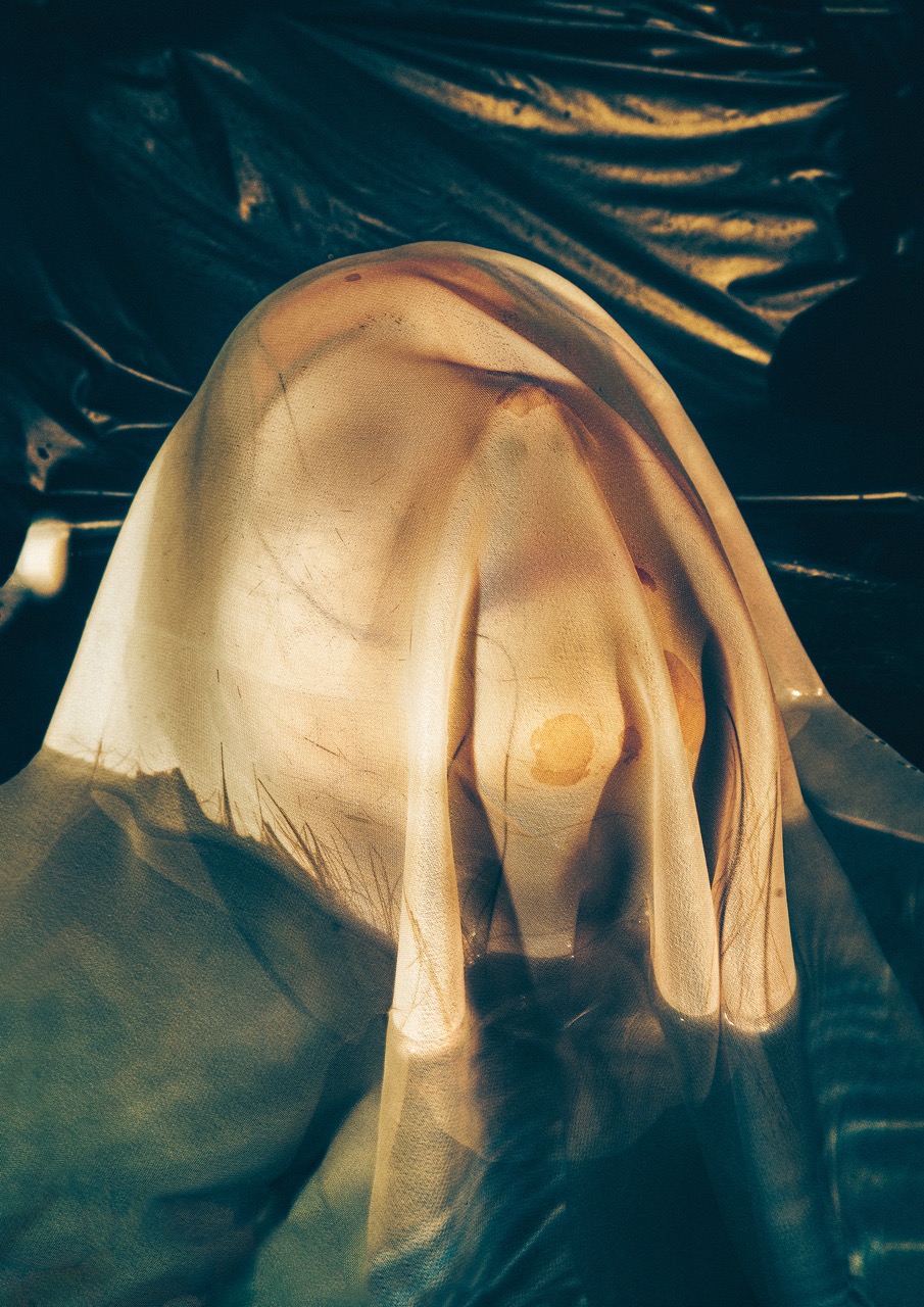A semi-transparent fabric drapes over a figure, revealing soft contours and textures beneath. The background features dark surfaces, enhancing the ethereal quality of the scene.