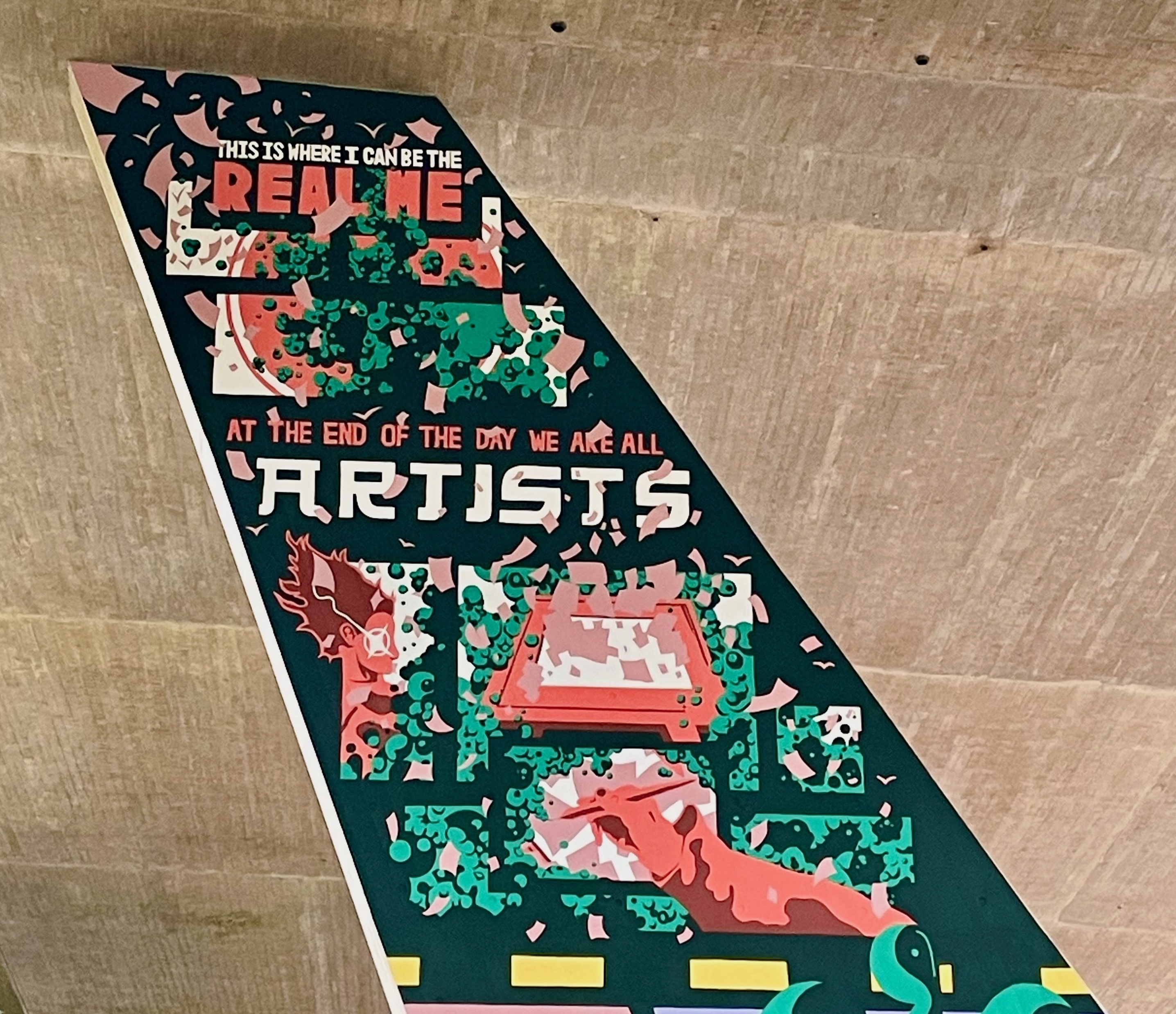 Part of a mural on a flyover pillar wall. Primarily black background featuring red and green abstract imagery and text reading "This is where I can be the real me" and "At the end of the day we are all artists".