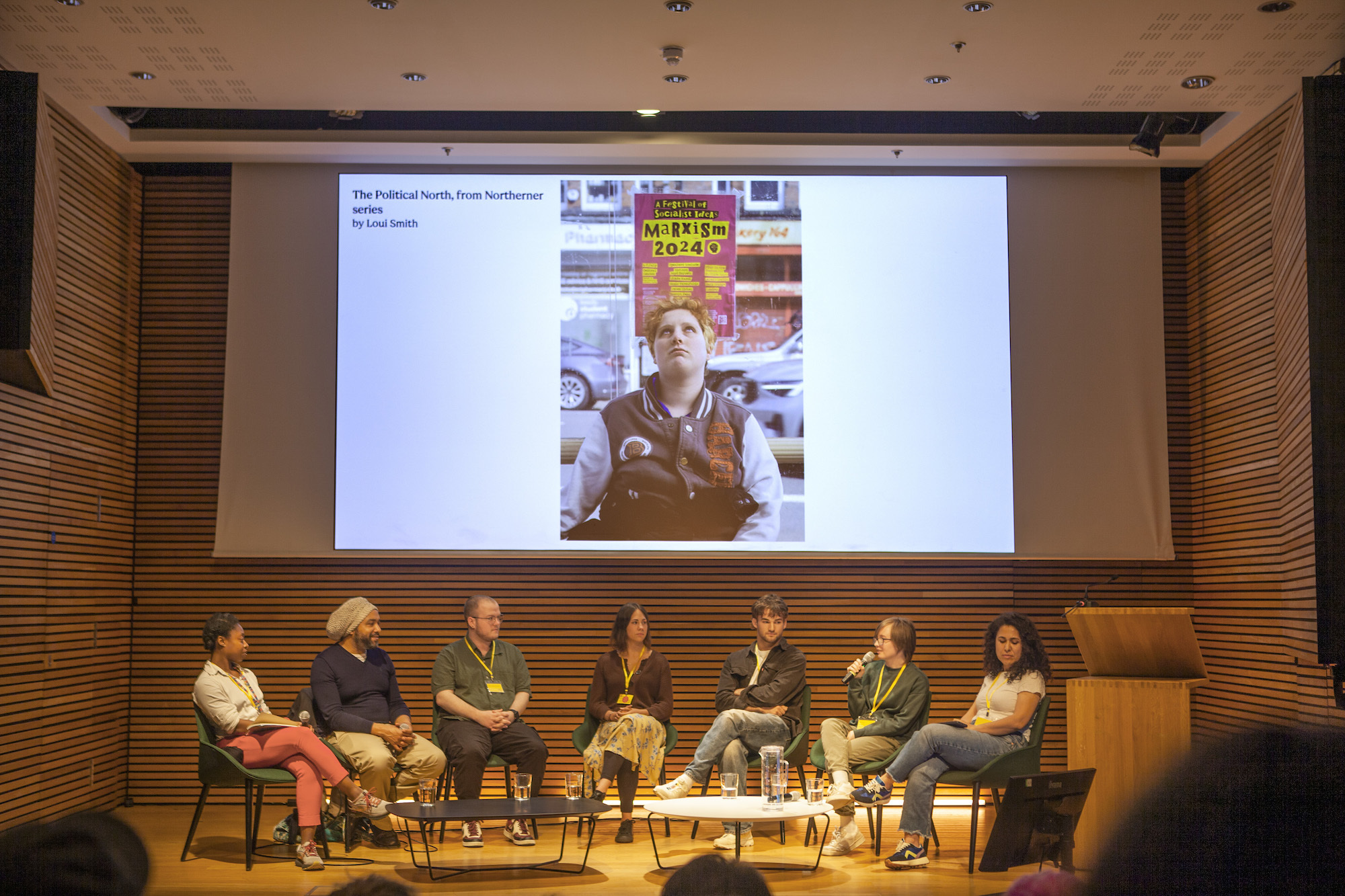 A group of people at a talk in a wood-wall seminar space with a projector screen showing photography.