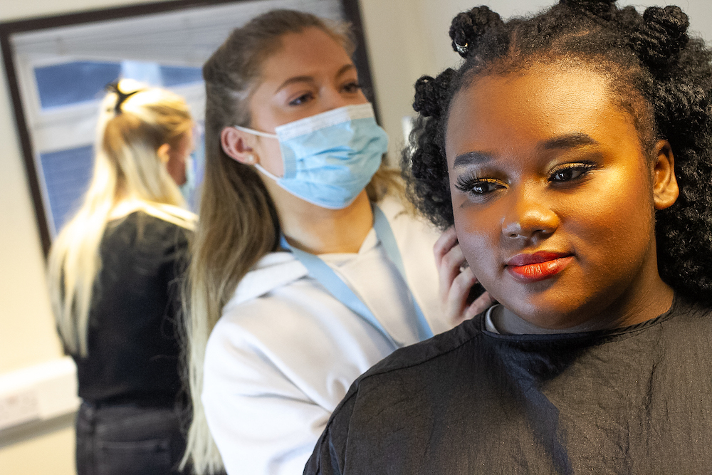 A make-up artist wearing a face mask adjusts an actor's hair from behind.