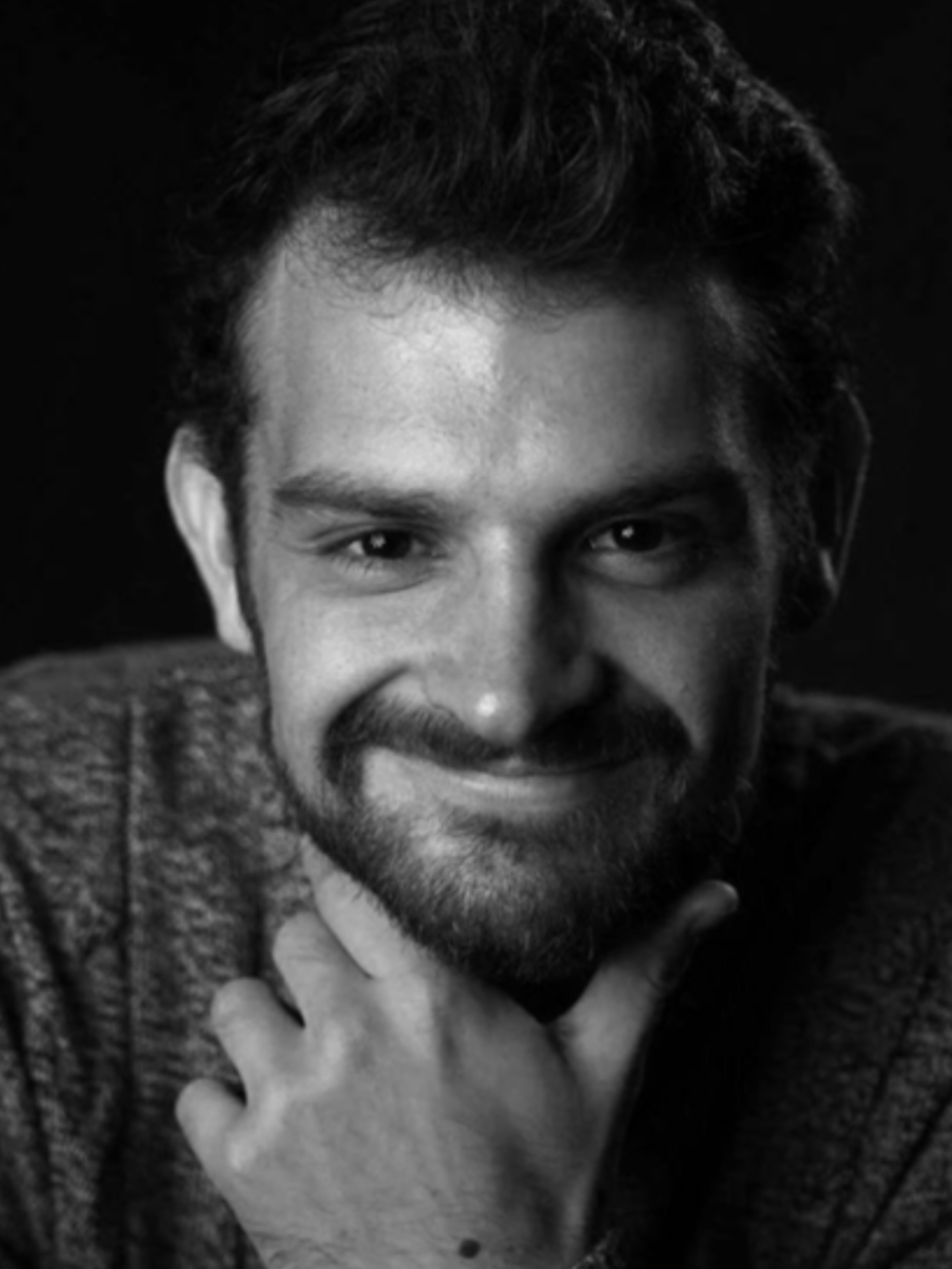 Black and white portrait of smiling man with beard and curly hair, hand positioned under chin, wearing knitted jumper.