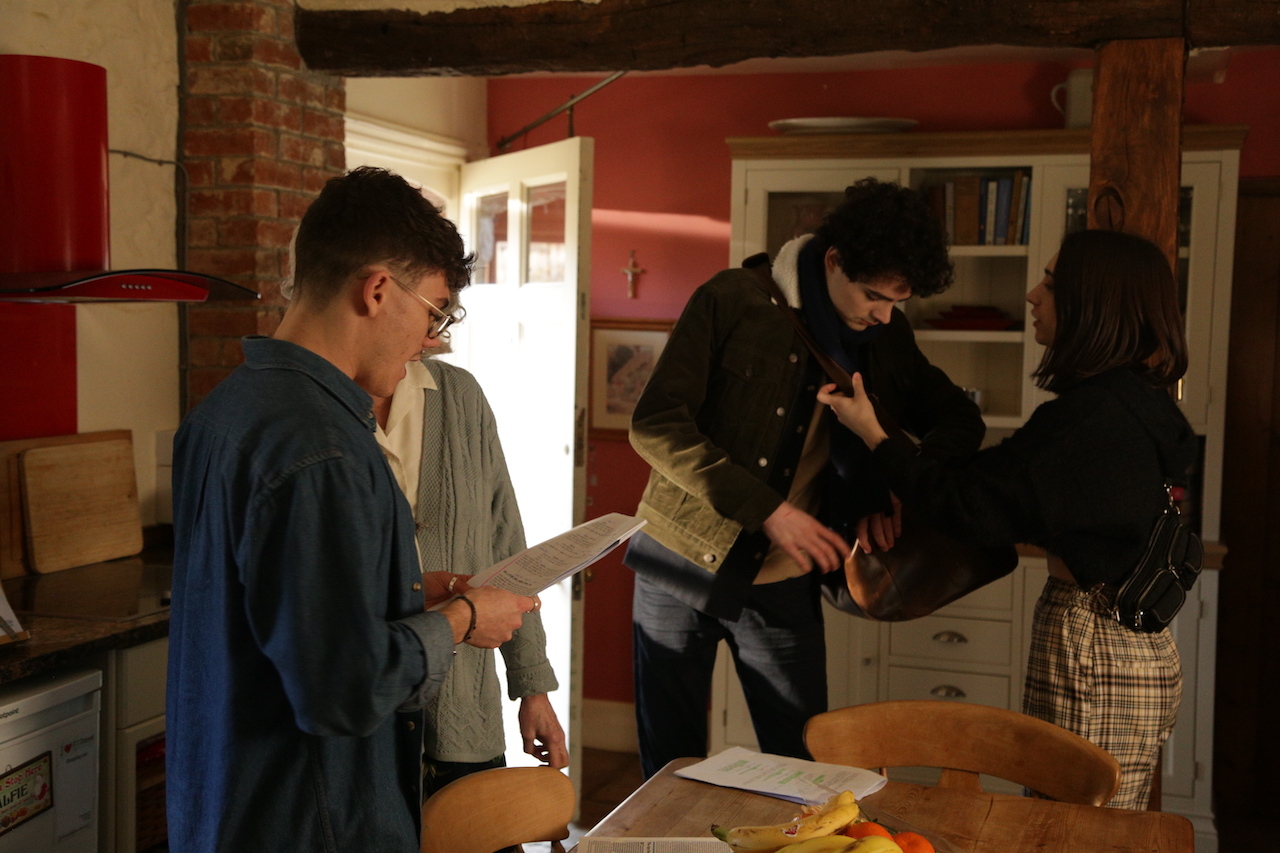 Three people in rustic kitchen with exposed beams, red walls, and wooden surfaces. Man with glasses holds paper whilst others examine documents on table.