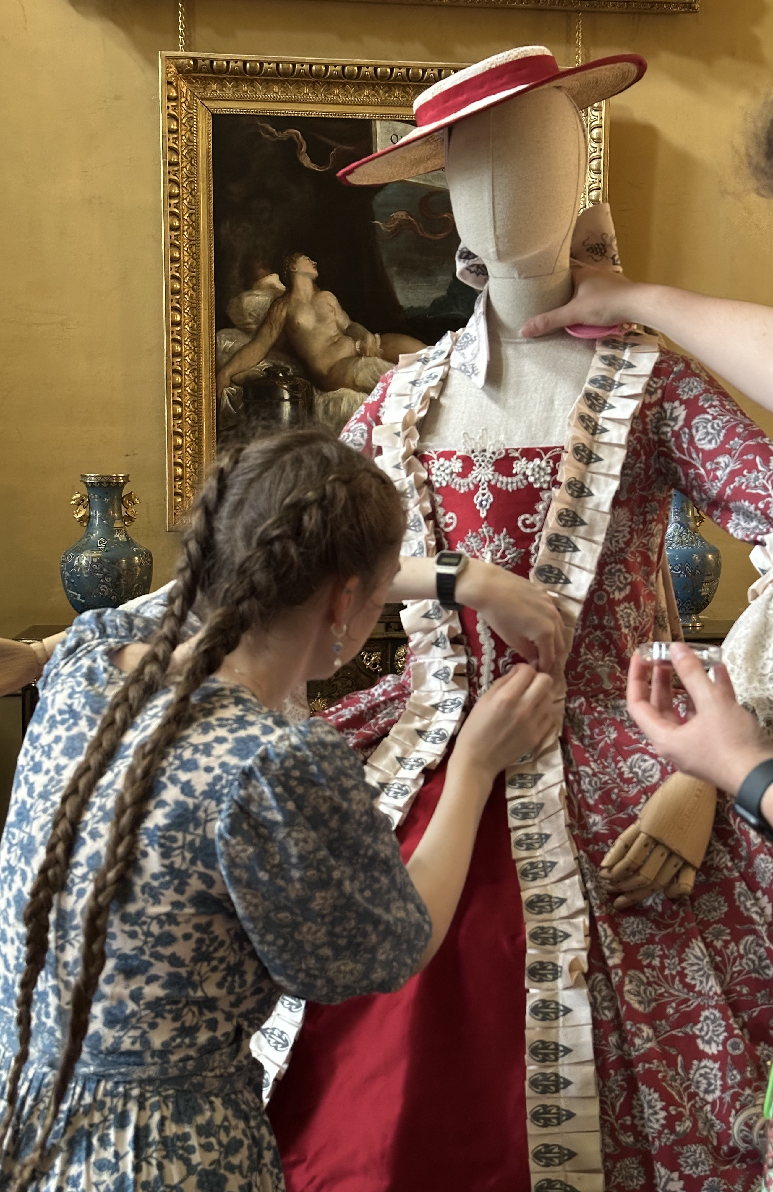 A woman adjusts the sleeve of a mannequin dressed in a patterned red dress and hat, with a framed painting and ceramic vases in the background.