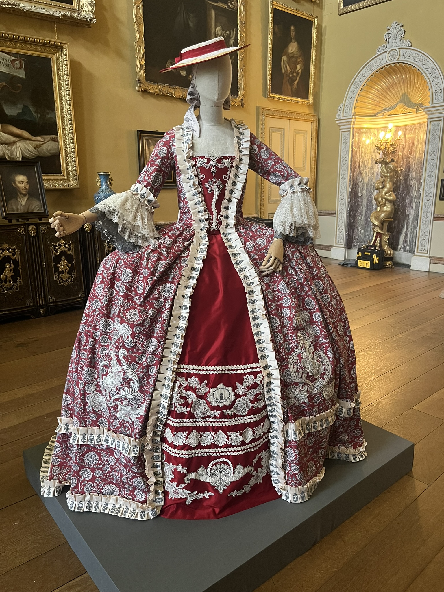 A mannequin displays an ornate red and pink Victorian-style gown with white lace trim, standing on a grey plinth in a museum room with framed portraits and an arched doorway.