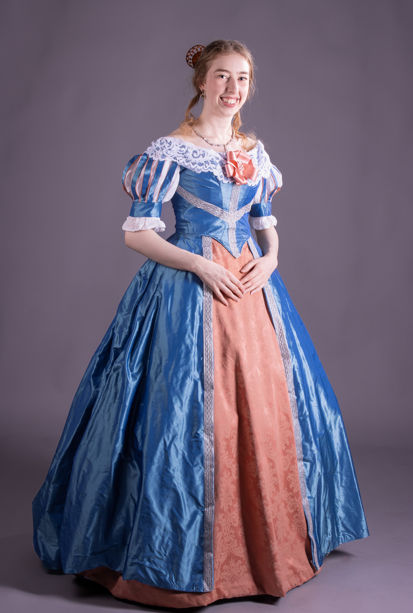 A young woman in a blue and peach historical-style gown poses against a plain grey backdrop.
