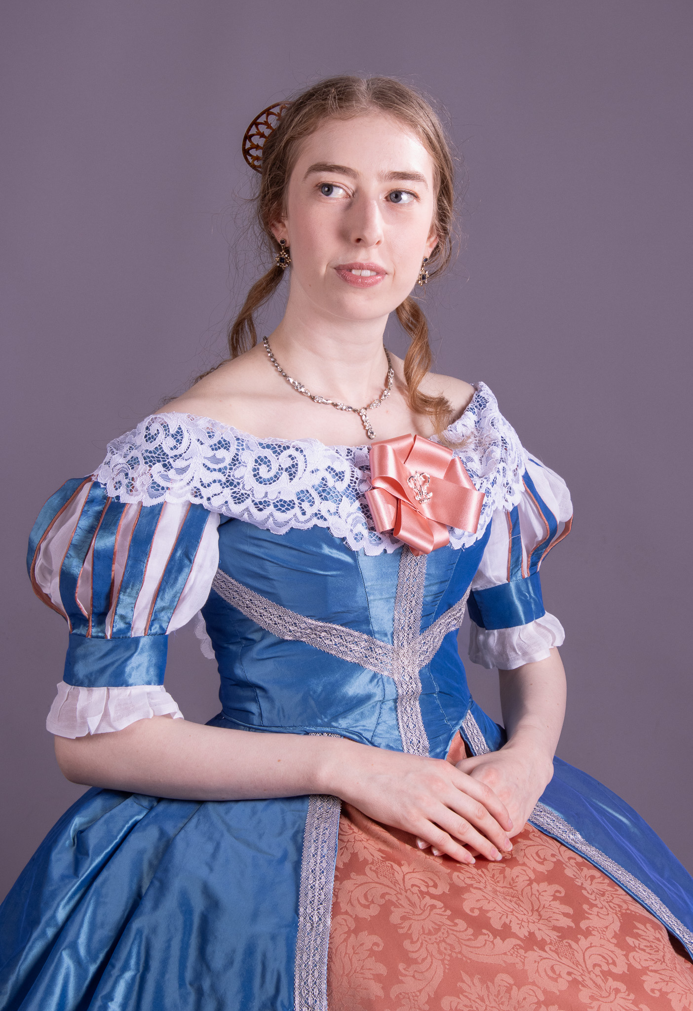 A young woman in a blue historical-style dress with lace detailing and a pink bow sits against a plain grey background.
