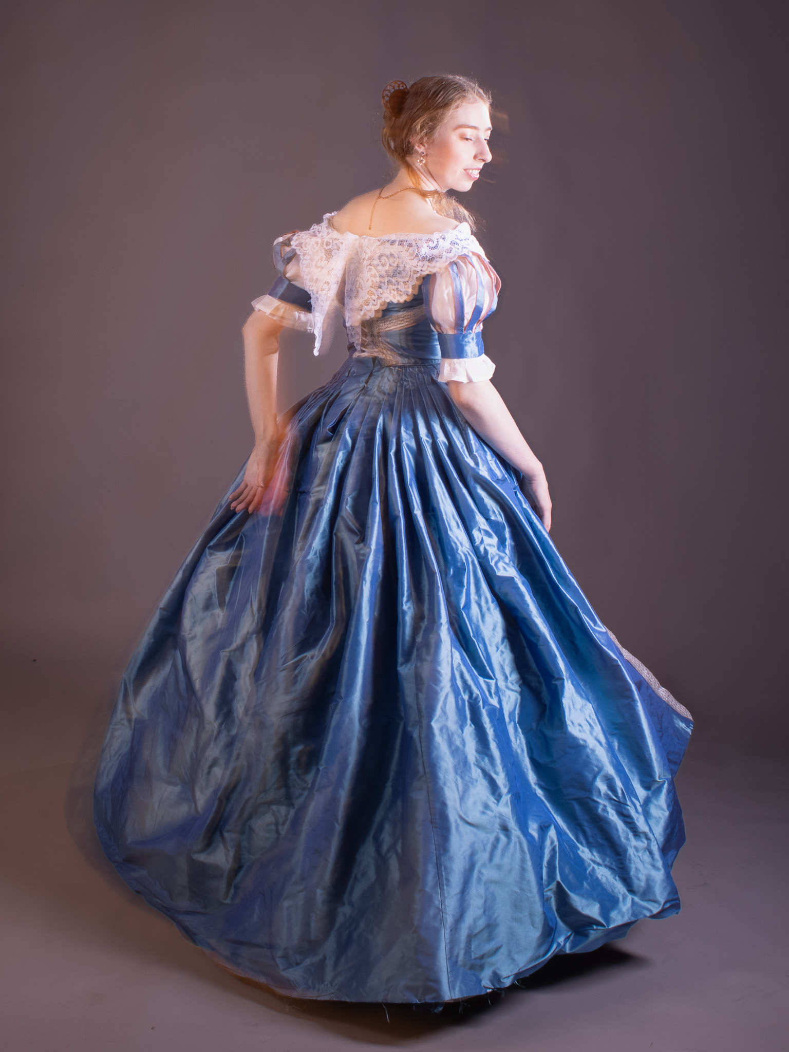 A woman in a blue Victorian-style gown with a white lace bodice stands against a plain grey studio backdrop.