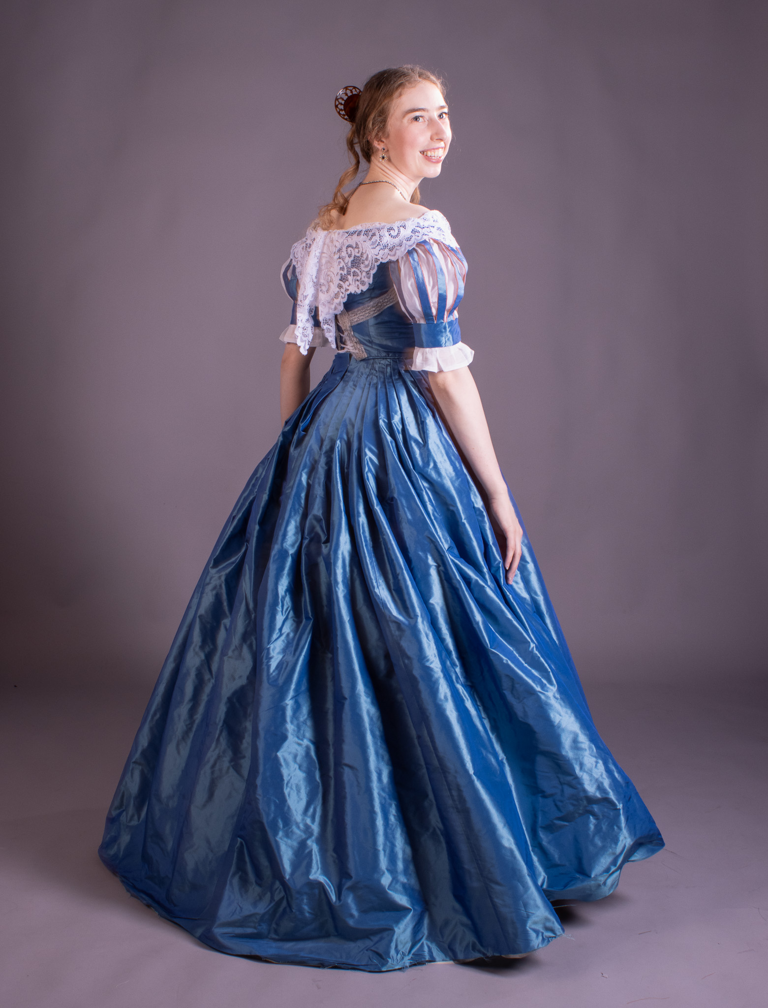 A young woman in a blue satin ball gown with lace trim, standing against a plain grey studio backdrop.