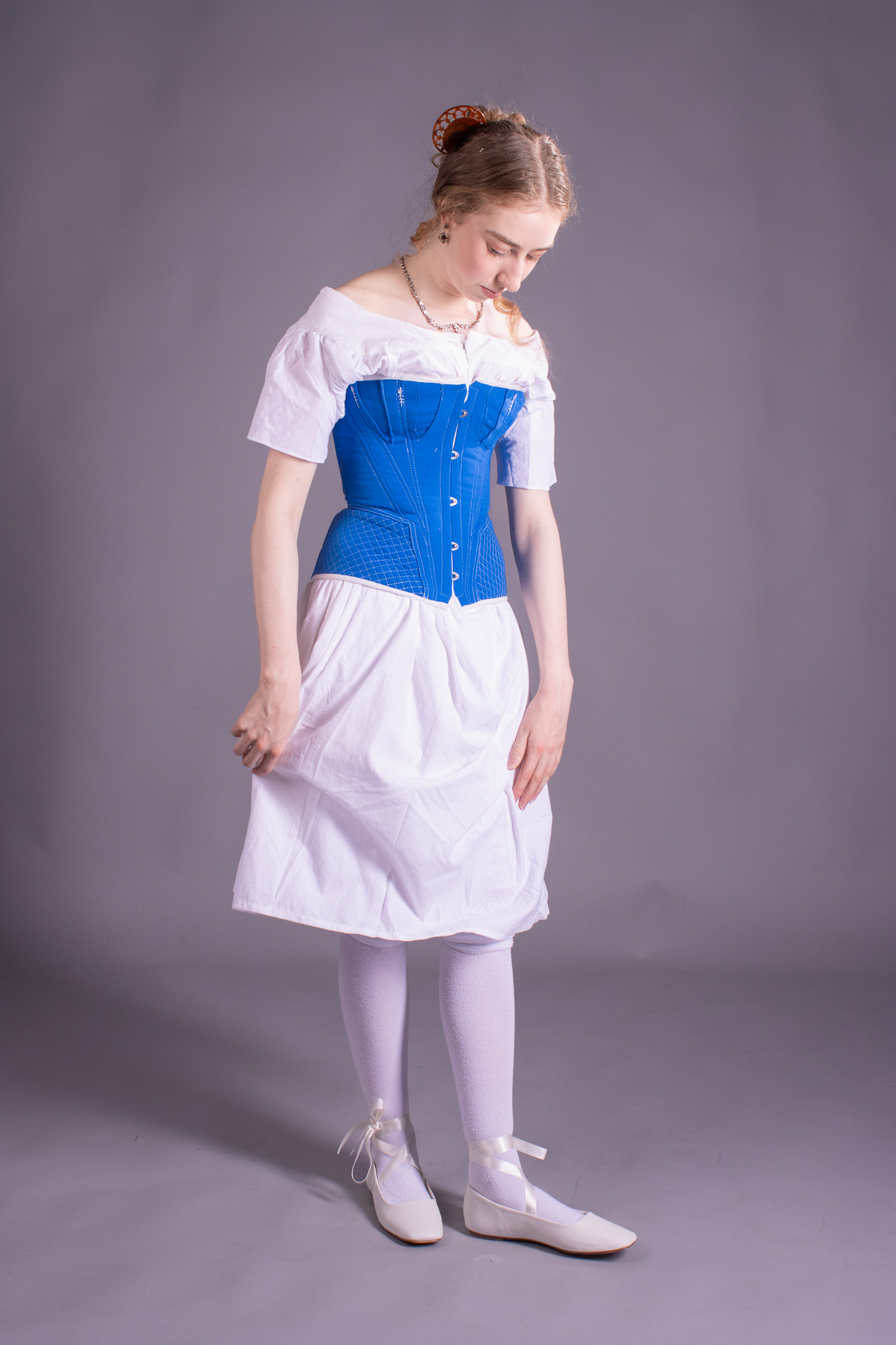 A young woman in a white dress, blue corset and white tights stands against a plain grey studio backdrop.