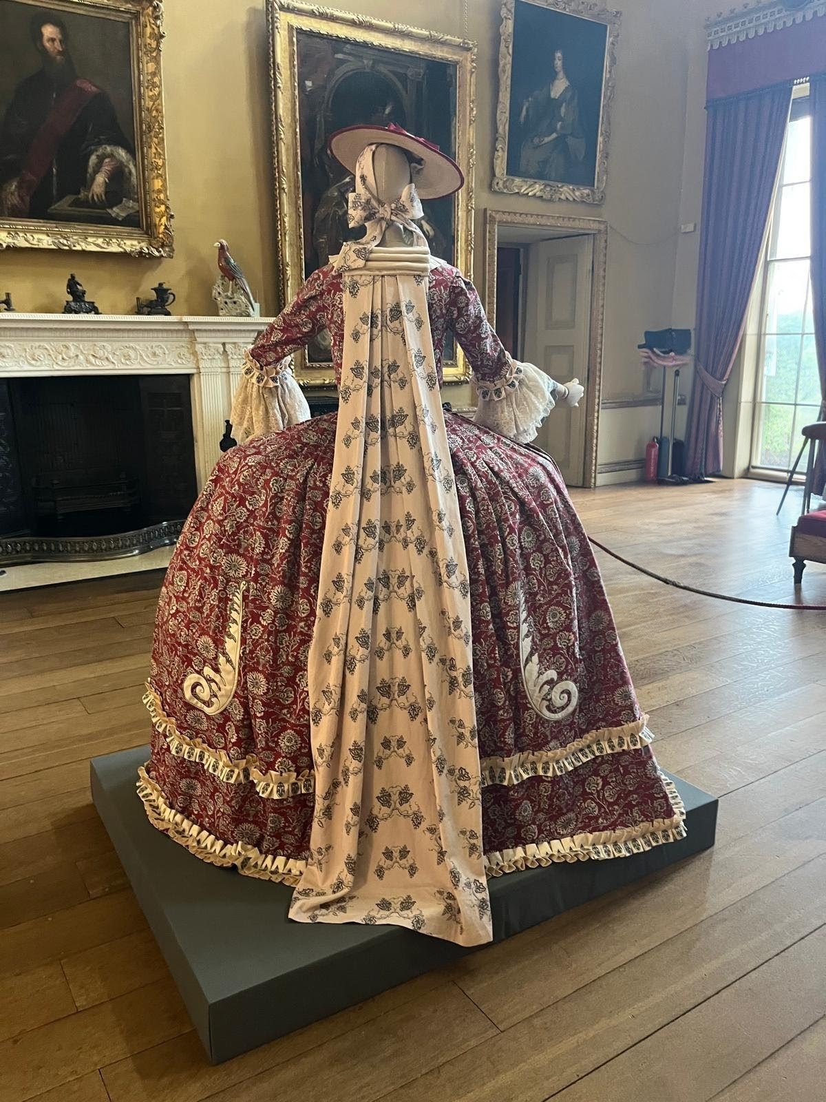 A historical mannequin in a patterned burgundy gown with a flowing cream floral train stands on a black plinth in a museum room, beside a fireplace and framed portraits.