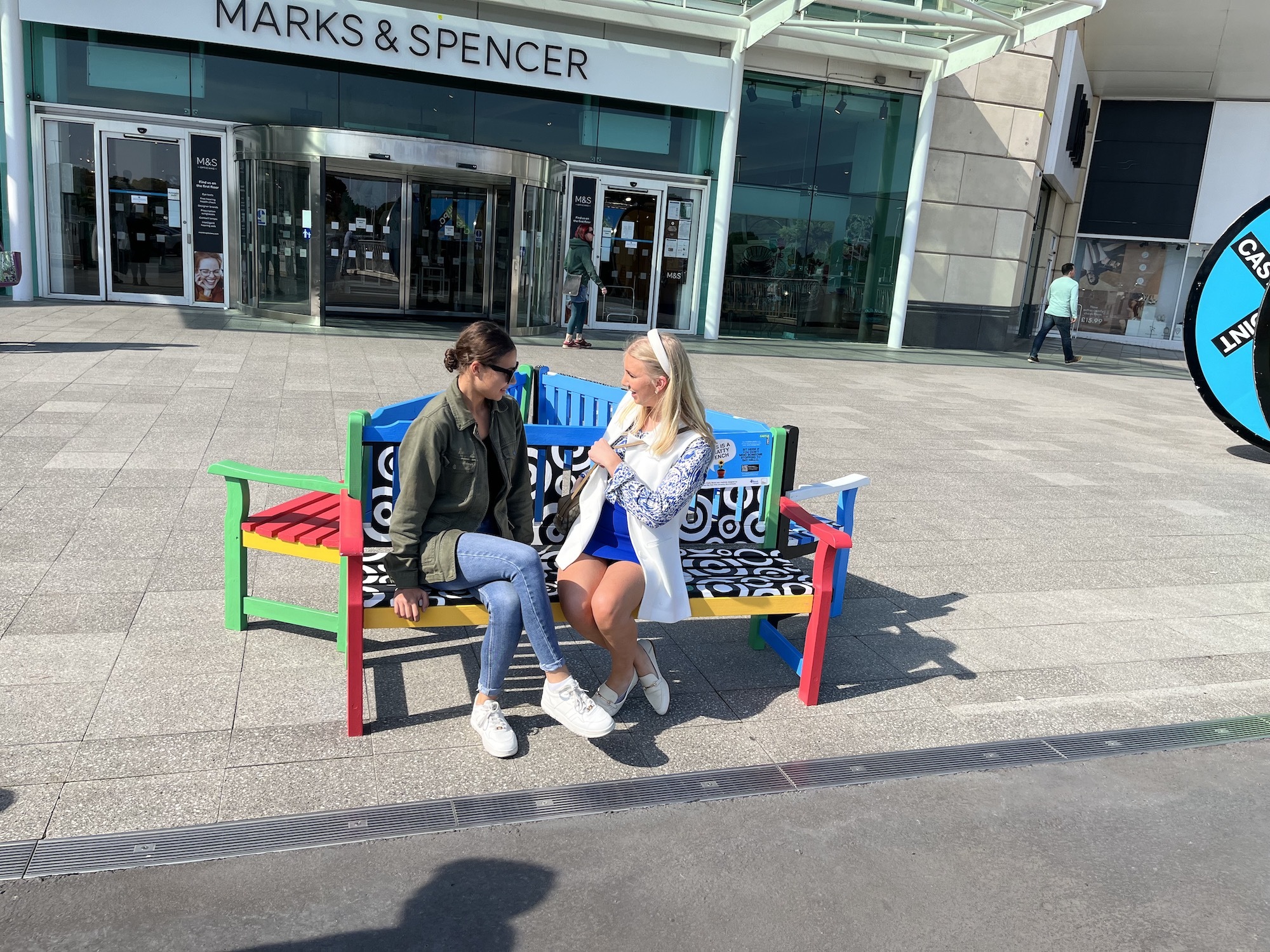 A BA (Hons) Illustration student sitting with a Castlepoint employee on one of the Chatty Benches in front of a Marks & Spencer shop.