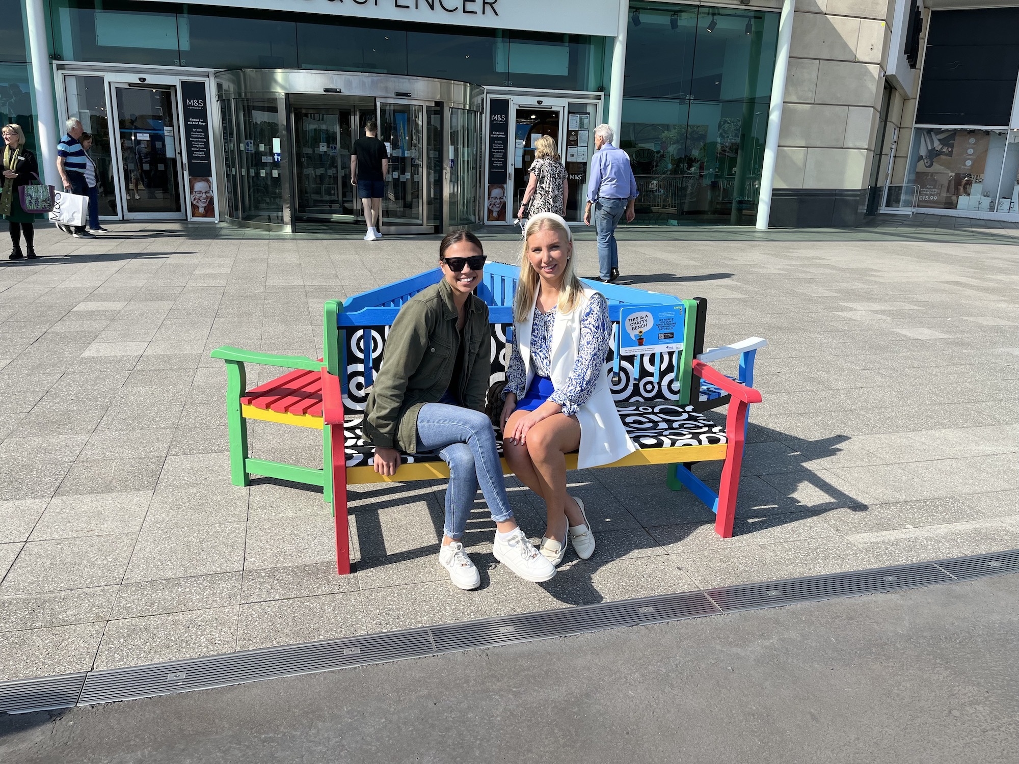 A BA (Hons) Illustration student sitting with a Castlepoint employee on one of the Chatty Benches in front of a Marks & Spencer shop. Both are looking at the camera and smiling.