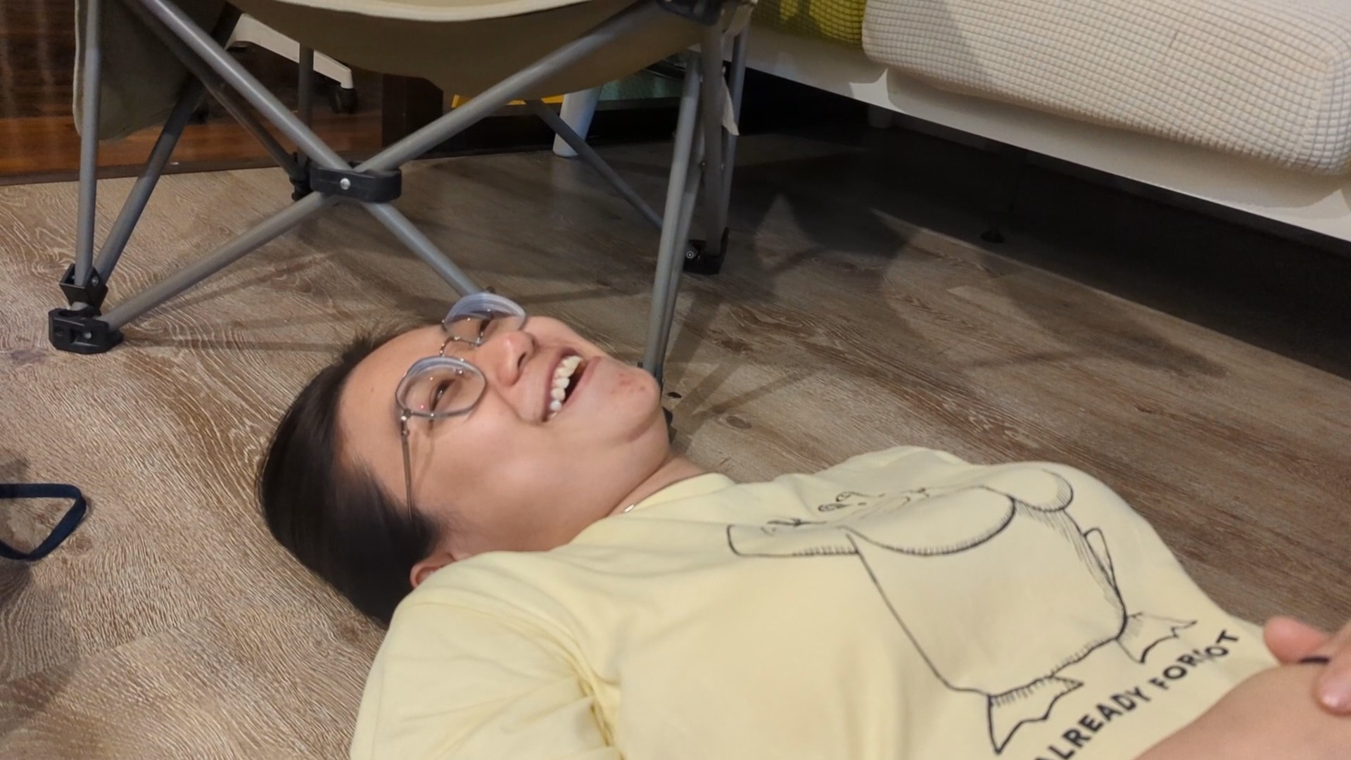 A woman wearing glasses lies on a wooden floor beneath a folding chair and sofa, with one hand resting on her stomach.