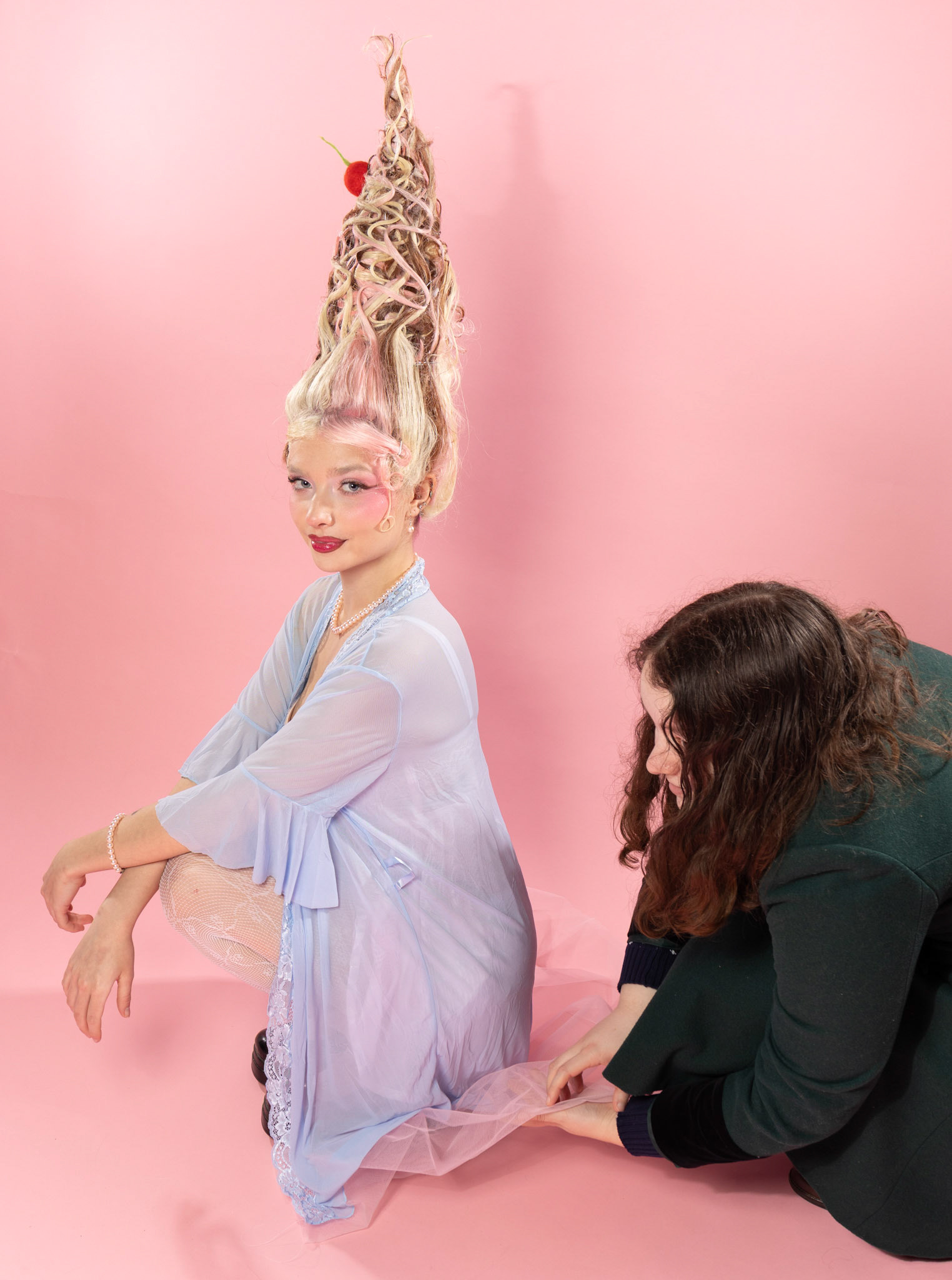 A woman with an elaborate high hairstyle adorned with a cherry, wearing a light blue outfit, poses on a pink background. Another person, dressed in a dark green outfit, adjusts the hem of her clothing from behind.