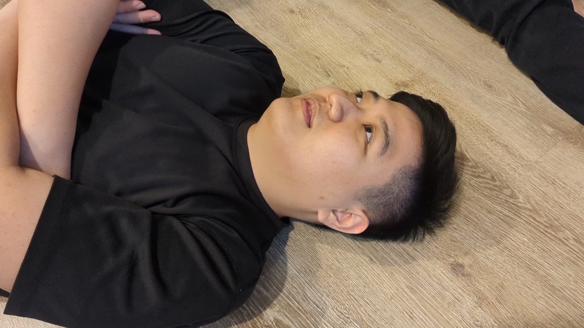 A man with short black hair lies on a wooden floor in a black T-shirt, with his arms crossed and another person’s leg partly visible nearby.