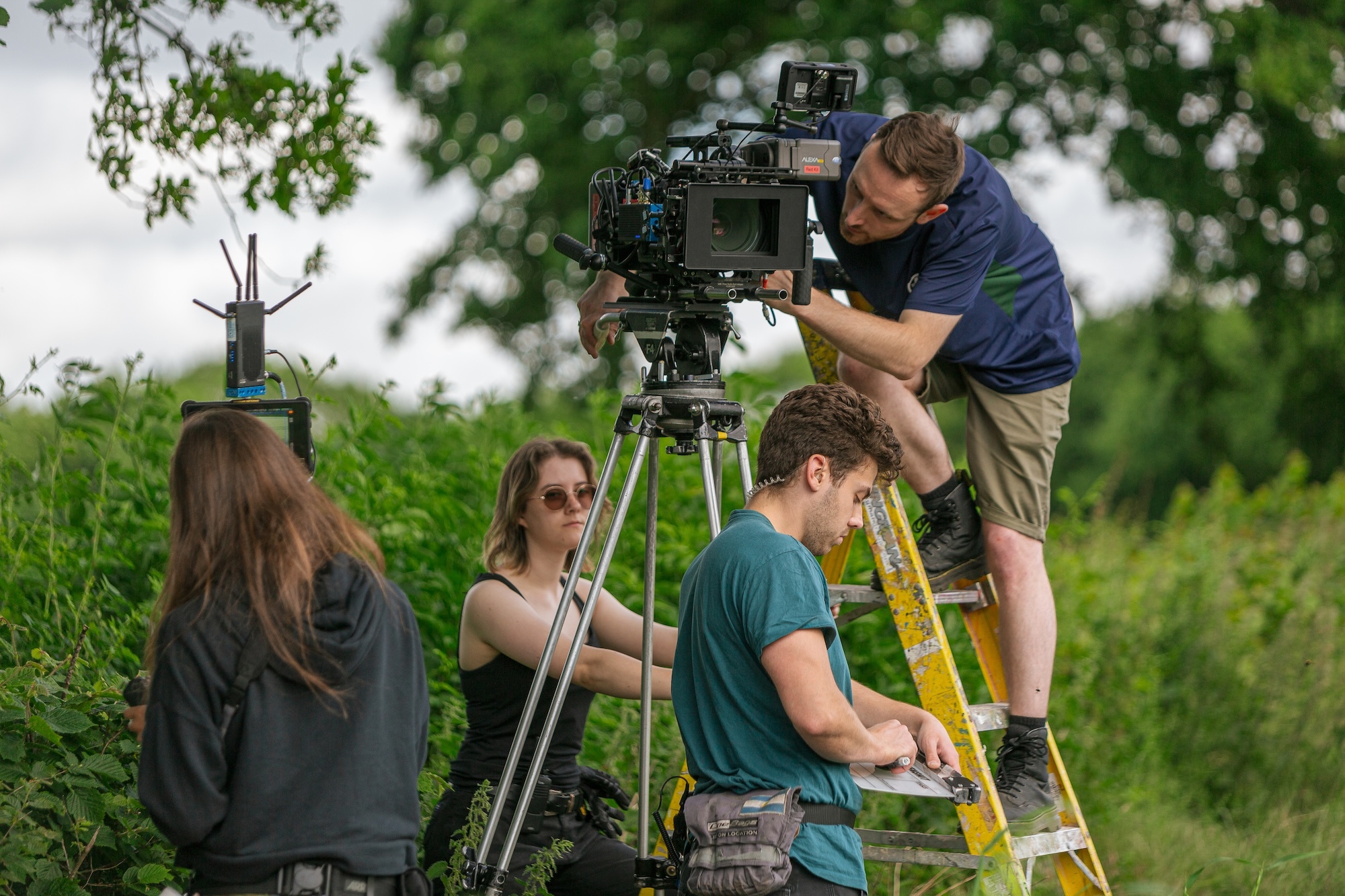 Four crew members on a location shoot for "Chickenshit". Two crew members are examining cameras, one standing on a ladder; the other two are in front of the tripod and behind the tripod. There are trees and other greenery in the background.