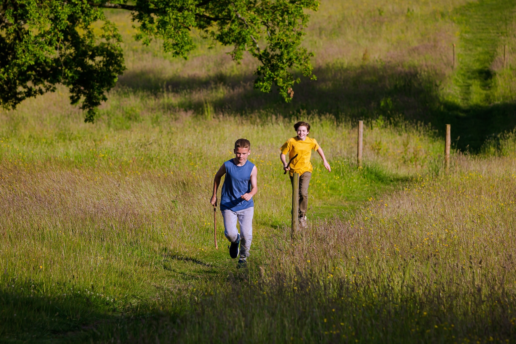Two young people running after one another in a field, both carrying large sticks.