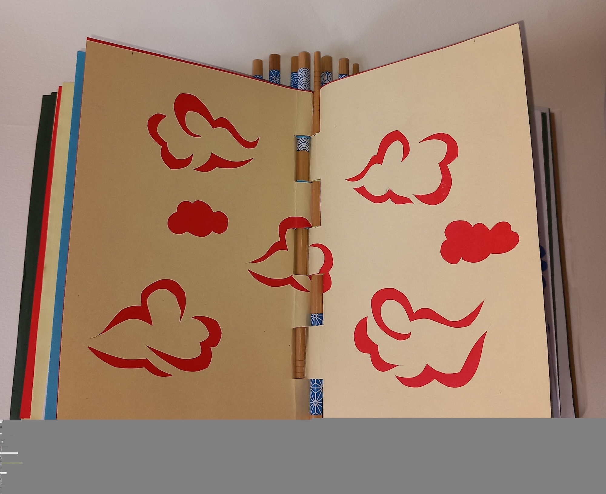 Open notebook with red cloud shapes drawn on cream pages, coloured pencils visible in spine, other notebooks stacked behind
