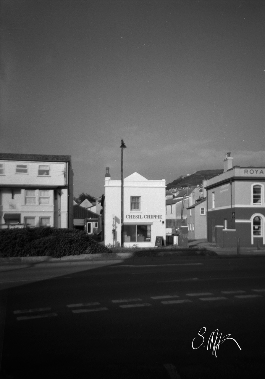 Black and white street scene with white shopfront marked "CHESIL CHIPPIE" between larger buildings, hills in background, cloudy sky.