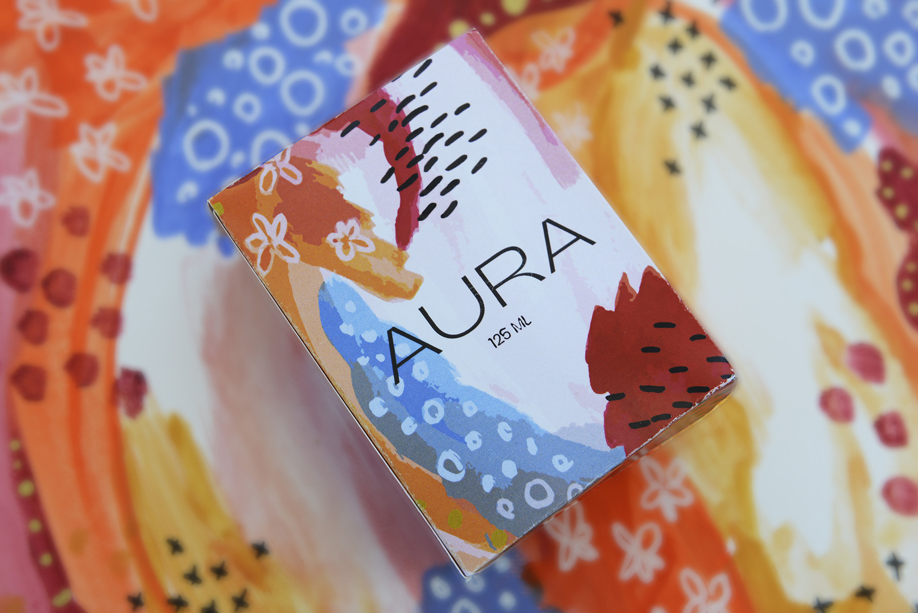 Colourful packaging box for a perfume called 'Aura', resting on a multi-coloured surface.