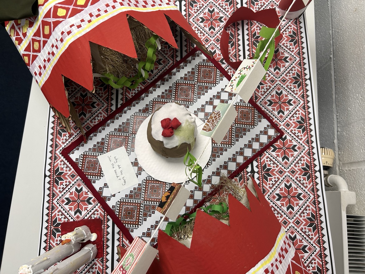 Colourful, textured red festive table with large red model cracker on either side, model Christmas pudding in the middle.