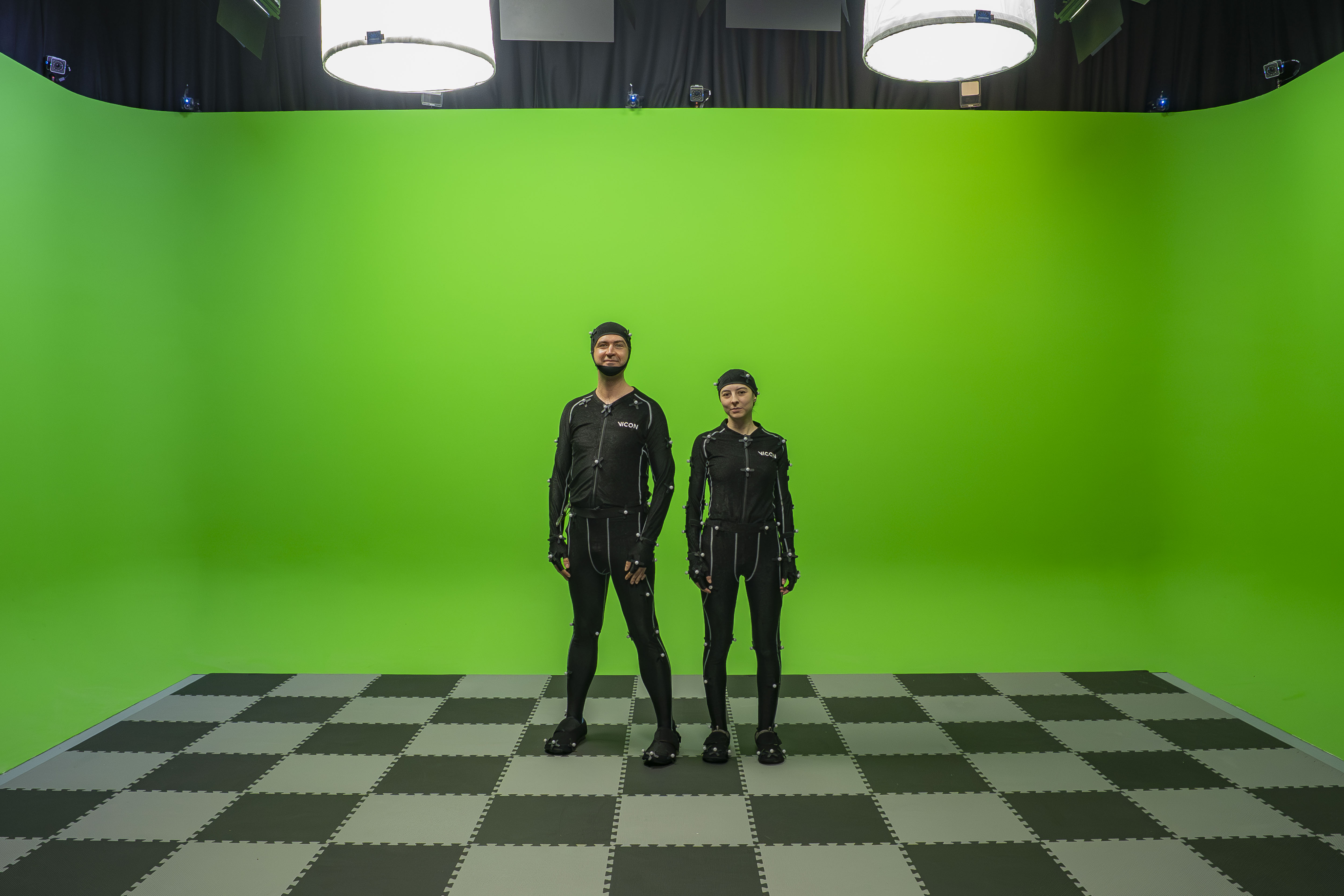 Two people wearing motion capture suits standing and posing in front of a Chroma-Key green surround.